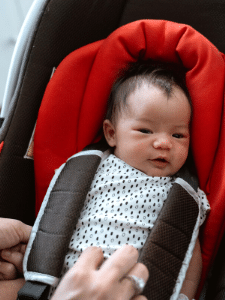 An infant smiles while being buckled into a car seat by a parent's hands