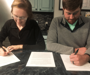 Photo is of a woman and man at a dining table signing contracts.