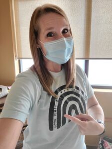 Photo of Jen wearing a COVID-protective mask and pointing to her t-shirt that says "Good Vibes Only"