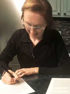 Photo is of a woman at a dining room table signing a contract.
